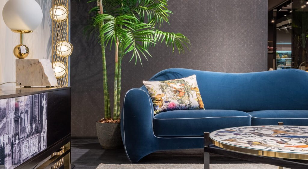 La imagen muestra una sala moderna y elegante, donde el protagonista es un sofá azul de diseño curvo acompañado por una mesa de centro artística y una lámpara decorativa de estilo contemporáneo. Todo está dispuesto con un equilibrio visual que transmite sofisticación y confort. A la izquierda del sofá, se destaca una Palma de bambú (Chamaedorea seifrizii), que aporta frescura y un toque tropical al ambiente. Su porte alto y esbelto complementa perfectamente la estética del espacio, demostrando cómo una planta bien elegida puede realzar la decoración de interiores con estilo natural.
